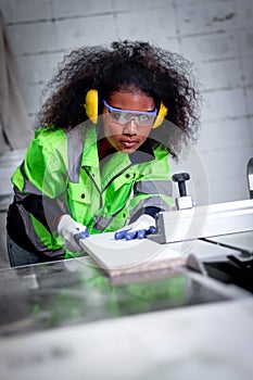 African industrial woman worker with curly hair wears safety vest and protective headphones, works with wood cutting machine,