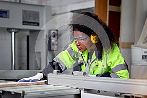 African industrial woman worker with curly hair wears safety vest and protective headphones, works with wood cutting machine,