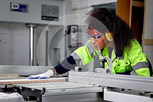 African industrial woman worker with curly hair wears safety vest and protective headphones, works with wood cutting machine,