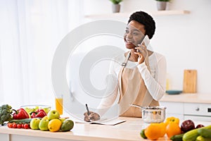 African Lady Cooking Talking On Cellphone Writing Recipes In Kitchen