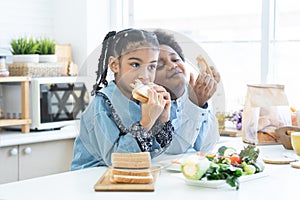 African little child girl eating homemade sandwiches in hands, standing in kitchen at home with brother, prepare food for