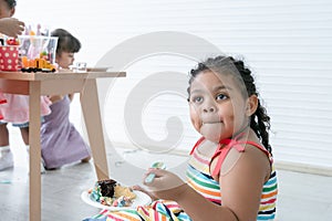 African little cute child girl sitting on floor, enjoy eating rainbow cake on birthday party by spoon with friends. Kids have fun