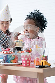 African little cute child girl use spoon enjoy eating big bite rainbow cake, sugar candies and jelly on birthday party with