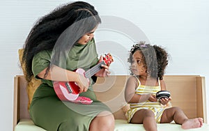 African little girl and mother playing ukulele together and smiling with happiness while doing leisure in living room at home on