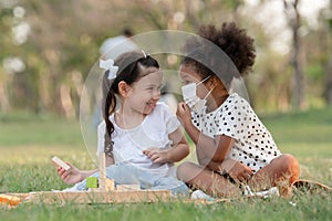 African little girl wear face mask talking to small Caucasian kid while sitting and playing wooden blocks toy in green park