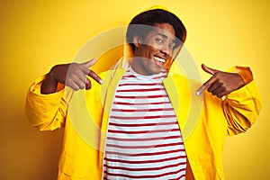 African man with afro hair wearing rain coat with hood standing over isolated yellow background looking confident with smile on