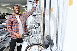 African man in a brown coat chooses a bathroom faucet in a construction supermarket, plumbing repairs in the house