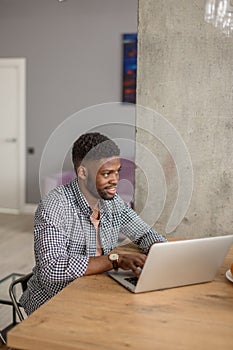 African man in casual wear sitting at table in front of open laptop at home