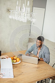 African man in casual wear sitting at table in front of open laptop at home
