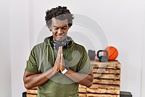 African man with curly hair wearing sportswear at the gym praying with hands together asking for forgiveness smiling confident