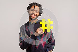 African man with dreadlocks and toothy smile on his face holding hashtag symbol, recommending popular topics, internet trends