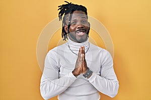 African man with dreadlocks wearing turtleneck sweater over yellow background praying with hands together asking for forgiveness