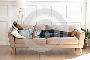 African man in headphones lying resting on couch with smartphone
