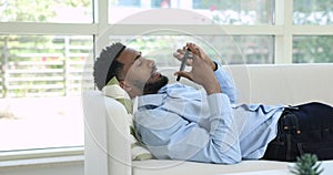 African man holding smartphone blab on speakerphone lying on sofa