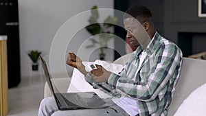 African man with laptop on knees, measuring body temperature with non-contact thermometer