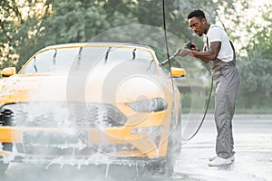 African man in protective overalls, washing his car manually with water high-pressure hose