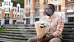 African man sitting lonely on city bench, holding flower bouquet, failed date