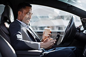 African man in suit drinking coffee and using mobile in car