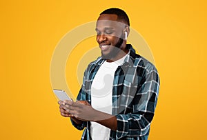 African Man Using Phone Wearing Earbuds Standing Over Yellow Background