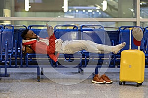 African man waiting for flight using smartphone. Black guy lying on bench near luggage in airport