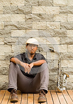 African man wearing sixpence hat and dark shirt sitting down leaning against wall, saxophone next to him