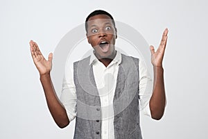 African man in white shirt and vest keeping mouth wide open, looking shocked after winning in lottery unexpectedly.