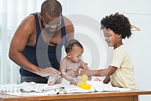 African middle aged father and child boy applying dusting powder on chest and leg of cute toddler baby girl at home after bathing