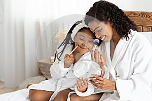 African mother and her little daughter in bathrobes making makeup on bed