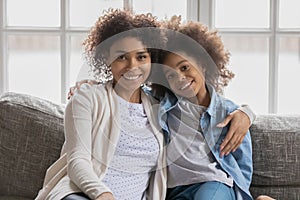 African mother hugs daughter sitting on couch looking at camera