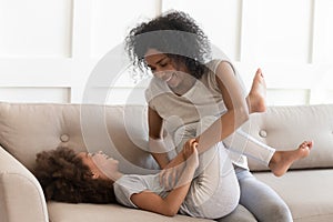 African mother tickles daughter sitting on couch have fun indoors