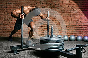 African muscular man pushing sled in cross training gym over brick wall