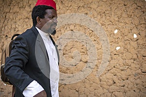 An African Older Man in Red Muslim Taqiyyah Fez Hat And Blazer on white Dress Moving a stick for lame people Near the