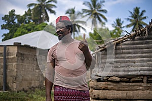 An African Older Man in Red Muslim Taqiyyah Fez Hat posing with a stick for lame people on Yard Near the Basic Hut with