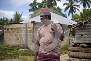 An African Older Man in Red Muslim Taqiyyah Fez Hat posing with a stick for lame people on Yard Near the Basic Hut with