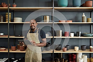 African potter with a tablet smiling by shelves in a ceramics workshop