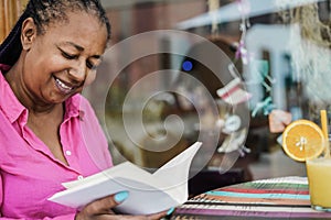 African senior woman reading a book outdoor at bar restaurant - Focus on face