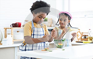 African siblings, boy in apron cooking, making mixing salad in bowl with little girl wear chef hat, holding pencil take note