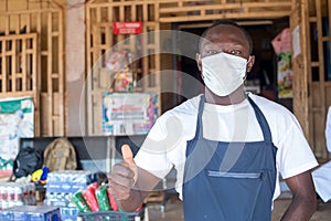 An african small business owner wearing a face mask in front of his store, gives a thumbs up gesture
