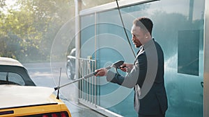 African stylish young focused man in business suit washing his modern sport car with a water gun on self-service washing