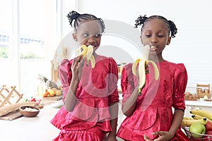 African twin girl sister with curly hair braid African hairstyle eating banana in kitchen. Happy smiling kid sibling eating fruit