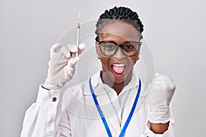 African woman with braids holding syringe screaming proud, celebrating victory and success very excited with raised arms