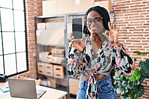 African woman with braids working at small business ecommerce showing smartphone screen doing ok sign with fingers, smiling