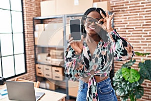 African woman with braids working at small business ecommerce showing smartphone screen smiling happy doing ok sign with hand on