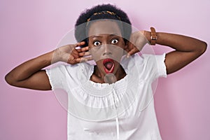 African woman with curly hair standing over pink background crazy and scared with hands on head, afraid and surprised of shock