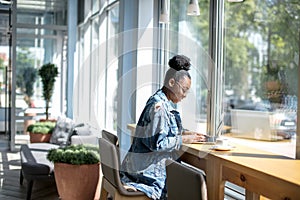 African woman in denim jacket sitting near the window and using laptop in cafe