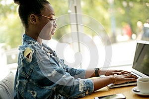 African woman in denim jacket sitting near the window and using laptop in cafe