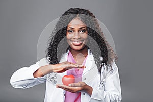 An African woman doctor in a white coat and with a stethoscope holds a red heart in her hands. cardiologist