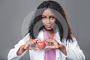 An African woman doctor in a white coat and with a stethoscope holds a red heart in her hands. cardiologist
