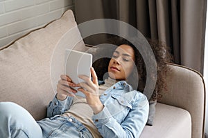 African woman holding digital tablet reading book lying on sofa at home.