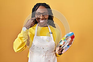 African woman holding painter palette pointing with hand finger to face and nose, smiling cheerful